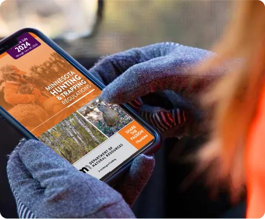 Gloved hands holding a phone that shows the Minnesota hunting and trapping regulations.